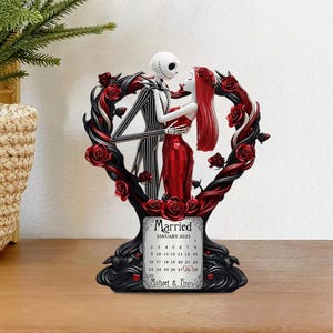 Puede incluir: Una figura decorativa con Jack Skellington y Sally de The Nightmare Before Christmas, abrazados dentro de un marco en forma de coraz&oacute;n con rosas rojas. La base incluye un calendario de enero de 2025, con la palabra "Married" y los nombres "Michael & Emma".