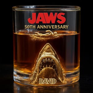 May include: A clear glass featuring a golden shark design and the name "DAVID". The glass is filled with a brown liquid and has the words "JAWS 50TH ANNIVERSARY" in red.