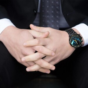 K&ouml;nnte beinhalten: Eine schwarze Armbanduhr mit rundem Zifferblatt und schwarzem Metallarmband. Das Zifferblatt der Uhr hat ein blaues und goldenes Design. Die Uhr wird am Handgelenk getragen, die Person tr&auml;gt einen schwarzen Anzug und ein wei&szlig;es Hemd.