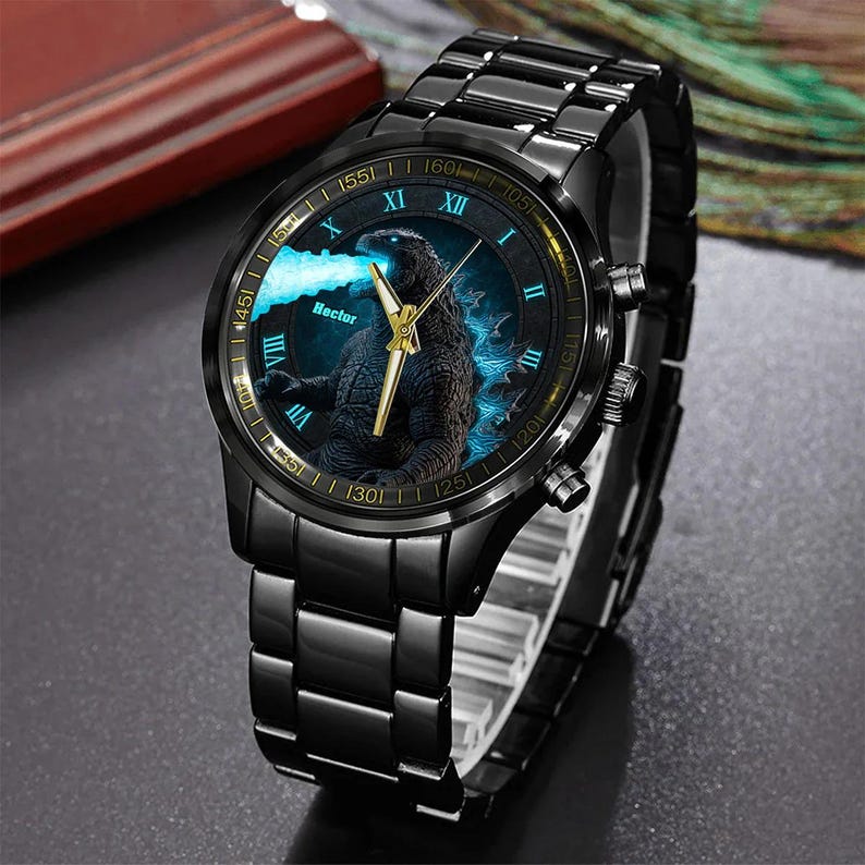 K&ouml;nnte beinhalten: Schwarze Armbanduhr mit einem Godzilla-Motiv auf dem Zifferblatt. Die Uhr hat ein schwarzes Metallarmband und goldfarbene Zeiger. Das Zifferblatt zeigt r&ouml;mische Ziffern und den Namen "Hector".