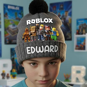 May include: A gray knit beanie featuring the word "ROBLOX" and the name "EDWARD" in white block letters. The beanie has a black pom-pom on top and a colorful graphic of ROBLOX characters. The beanie is designed for children.