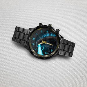 K&ouml;nnte beinhalten: Schwarze Armbanduhr mit Godzilla-Design auf dem Zifferblatt. Die Uhr hat ein schwarzes Metallarmband und ein rundes Zifferblatt mit goldenen Zeigern und r&ouml;mischen Ziffern. Das Zifferblatt hat ein blaues und schwarzes Farbschema.
