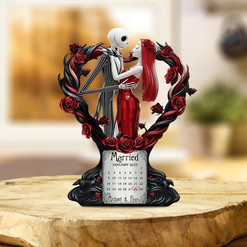 Puede incluir: Figura decorativa que representa a Jack y Sally de Pesadilla antes de Navidad, abraz&aacute;ndose dentro de un marco en forma de coraz&oacute;n adornado con rosas rojas. La base incluye un calendario para enero de 2025, con los nombres "Michael & Emma" debajo.