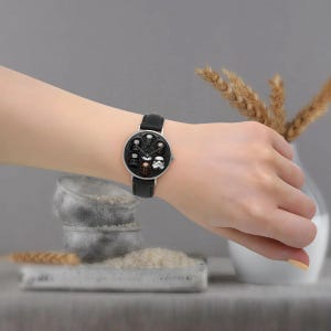 May include: A black leather watch with a silver-colored face featuring Star Wars character designs. The watch has a black leather band and is worn on a wrist.