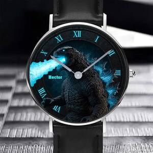 K&ouml;nnte beinhalten: Schwarze Armbanduhr mit silberner L&uuml;nette und schwarzem Lederarmband. Das Zifferblatt zeigt eine Godzilla-Grafik mit blauen Akzenten und dem Namen "Hector". R&ouml;mische Ziffern kennzeichnen die Stunden.