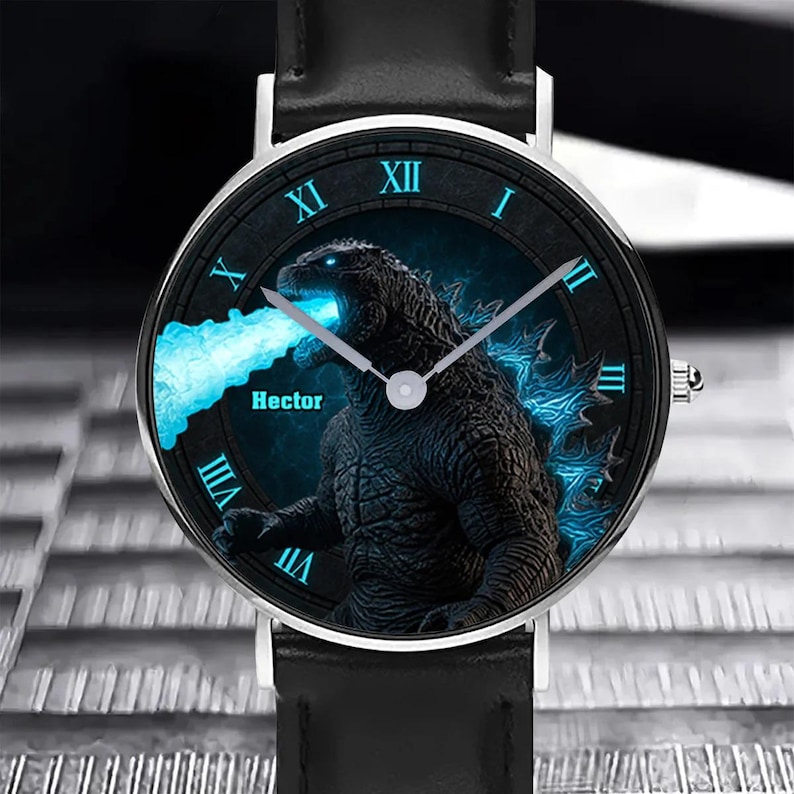 K&ouml;nnte beinhalten: Schwarze Armbanduhr mit silberner L&uuml;nette und schwarzem Lederarmband. Das Zifferblatt zeigt eine Godzilla-Grafik mit blauen Akzenten und dem Namen "Hector". R&ouml;mische Ziffern kennzeichnen die Stunden.