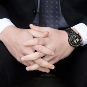 May include: Black wristwatch with a round face and a black metal band. The watch face features a gold-colored design with a white graphic. The watch is worn on a wrist, with the person wearing a black suit and white shirt.
