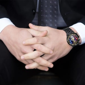 May include: Black wristwatch with a round face featuring a Star Wars design. The watch has a black metal band and a detailed face with a character and the text "MAY THE FORCE BE WITH YOU". The watch is on a person's wrist.