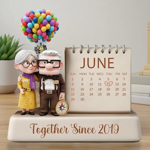 Peut inclure: Figurine représentant un couple enlacé, avec un calendrier affichant "Juin" et une date en surbrillance. Une boussole et un groupe de ballons colorés avec une maison sont également présents. La base indique "Together Since 2019".