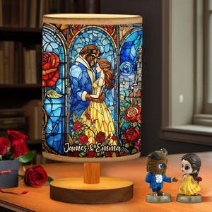 Op de afbeelding: Decoratieve lamp met een glas-in-lood ontwerp met de personages Belle en het Beest. De lamp heeft een houten voet en een cilindrische lampenkap met de tekst "James & Emma". Er zijn ook twee beeldjes van de personages aanwezig.