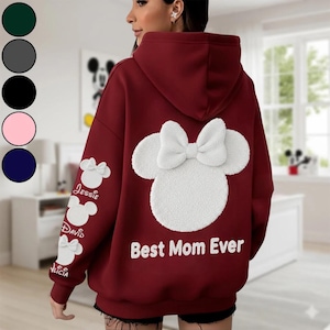 Könnte beinhalten: Burgunderfarbener Kapuzenpullover mit weißem Minnie-Maus-Design auf dem Rücken und den Ärmeln. Das Design umfasst eine große Schleife und den Schriftzug "Best Mom Ever". Die Ärmel zeigen Minnie-Maus-Designs mit Namen. Erhältlich in mehreren Farben.