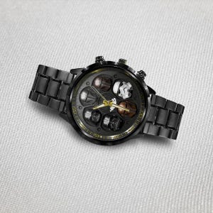 May include: Black wristwatch with a round face featuring various character helmets. The watch has a black metal band and a gold-colored bezel with markings. The watch face includes gold-colored hands and markers.