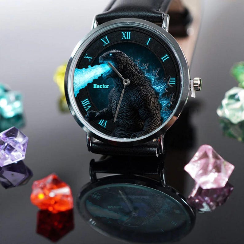 K&ouml;nnte beinhalten: Eine schwarze Armbanduhr mit silberner L&uuml;nette und schwarzem Lederarmband. Das Zifferblatt zeigt eine Godzilla-Grafik mit blauen Akzenten und dem Namen "Hector". Die Stunden sind in r&ouml;mischen Ziffern angegeben.