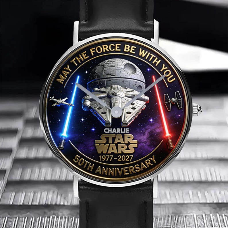 May include: A Star Wars themed watch with a black leather band. The watch face features the Death Star, Millennium Falcon, and lightsabers against a starry background. Text on the watch reads "May the Force Be With You" and "Star Wars 50th Anniversary".