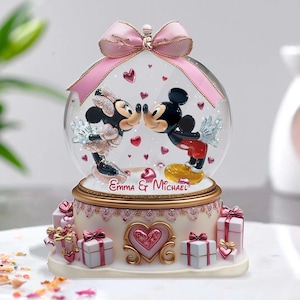 May include: A pink and white snow globe with Mickey and Minnie Mouse kissing inside. The base of the snow globe is decorated with pink and white gift boxes and a heart. The snow globe is personalised with the names "Emma & Michael".