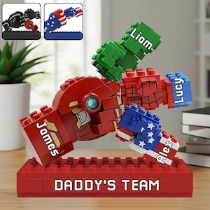 Może przedstawiać: Czerwona rzeźba w stylu Lego z napisem "DADDY'S TEAM" na podstawie. Rzeźba przedstawia czerwoną głowę i ramiona, z zielonymi, niebieskimi i czerwonymi pięściami. Imiona są wydrukowane na pięściach. To spersonalizowany prezent.