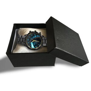 K&ouml;nnte beinhalten: Eine schwarze Armbanduhr mit blauem und schwarzem Zifferblatt mit Wasserfalldesign, pr&auml;sentiert in einer schwarzen Geschenkbox. Die Uhr hat ein schwarzes Metallarmband und goldfarbene Akzente.