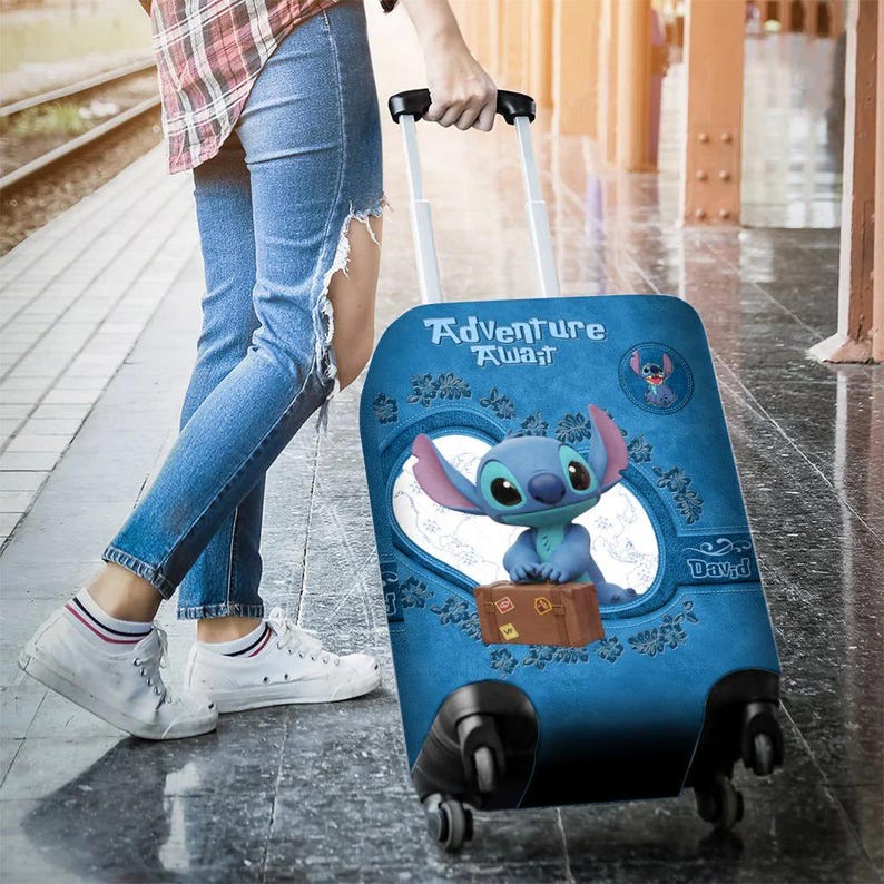 Puede incluir: Maleta con ruedas azul con el personaje Stitch de Lilo & Stitch. La maleta tiene las palabras "Adventure Awaits" y un peque&ntilde;o gr&aacute;fico de maleta marr&oacute;n. La maleta es tirada por una persona que lleva vaqueros azules y zapatillas blancas.