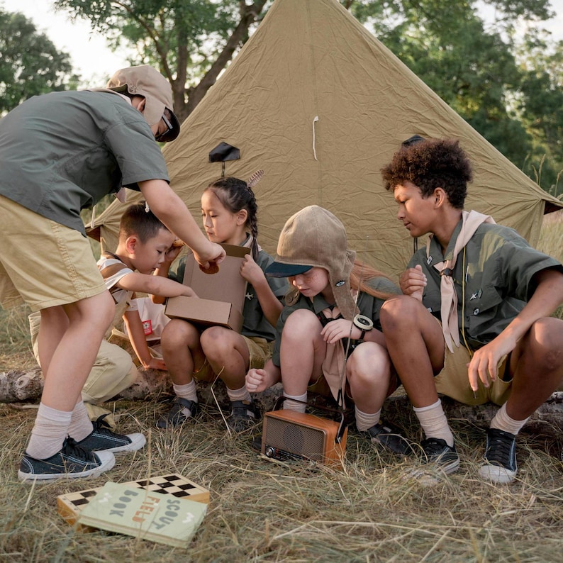 Peut inclure: Un groupe d'enfants sont rassembl&eacute;s autour d'une tente brune, jouant avec une bo&icirc;te en carton et une radio vintage. Ils portent des uniformes de scouts et profitent de leur temps en plein air.