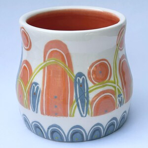 May include: A ceramic mug with a white base featuring hand-painted abstract designs in peach, light blue, and lime green. The exterior displays rounded shapes, vertical pillars, and a scalloped pattern at the base, while the interior is peach.