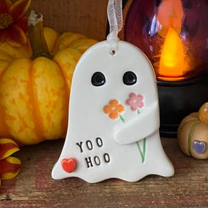 May include: A white ceramic ghost ornament featuring two black eyes, a small red heart, and the text YOO HOO. The ghost is holding a small orange flower and a pink flower with green stems. A white ribbon is attached to the top for hanging.