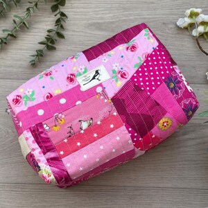 May include: A patchwork pouch in shades of pink, with a quilted design. The bag features a mix of floral, polka dot, and striped patterns. A small label with a bird logo is sewn on the front. The bag is likely handmade.