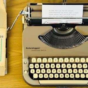 May include: A beige Scheidegger Princess-Matic typewriter with a white paper insert. The paper insert has German text that reads: "De kann viel tippen, mit der soliden Schreibmaschine, an besten immer 2 Blatt Papier nebeneinander, weil die Balken relativ hart ist.  Der Finger kann beim soliden..."