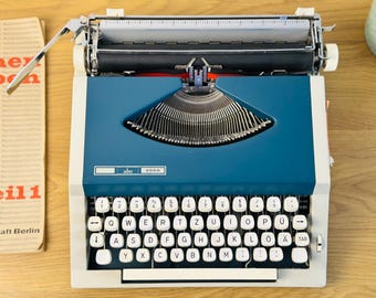 Może przedstawiać: Zabytkowa maszyna do pisania ABC 2000 z niebiesko-białą obudową. Klawiatura ma białe klawisze z czarnymi literami. Po lewej stronie znajduje się kartka z tekstem. Maszyna do pisania stoi na drewnianej powierzchni.