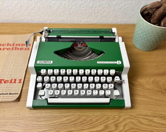 May include: A vintage Olympia Traveller de Luxe typewriter in green and white. The typewriter has white keys with black lettering and a chrome carriage. The image also shows a document and a potted plant.