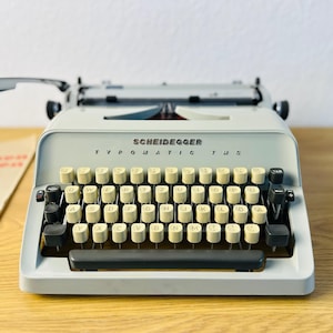 May include: A vintage Scheidegger Typomatic TM5 typewriter in light gray with cream-colored keys. The typewriter sits on a wooden surface, with a printed document to the left. The keys are arranged in a QWERTY layout.