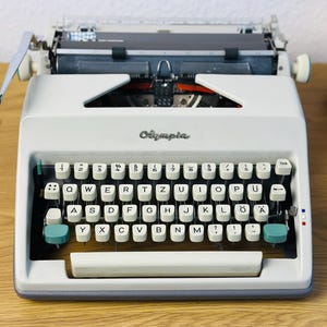 Puede incluir: Una máquina de escribir Olympia vintage blanca con un teclado verde y blanco. La máquina de escribir tiene una palanca de retorno de carro negra y una cinta negra y blanca.