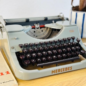 Könnte beinhalten: Eine vintage hellblaue Schreibmaschine mit einer schwarzen Tastatur und dem Wort "MERCEDES" auf der Vorderseite. Die Schreibmaschine hat einen schwarzen Wagenrücklaufhebel und eine schwarze Walze.