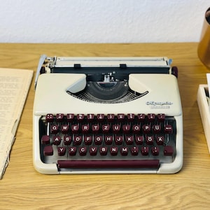 Könnte beinhalten: Eine weiße Vintage-Schreibmaschine mit einer burgunderfarbenen Tastatur und dem Wort "Olympia" auf der Vorderseite. Die Schreibmaschine steht auf einer Holzoberfläche.