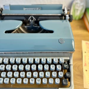 May include: A light blue vintage typewriter with a black keyboard and silver accents. The keys have German lettering.