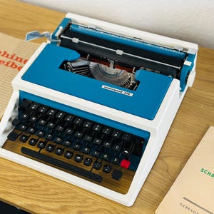 Puede incluir: Una máquina de escribir vintage Underwood 315 blanca y azul con un teclado negro y un botón rojo. La máquina de escribir está sobre una superficie de madera.