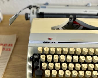 Może przedstawiać: Zabytkowa maszyna do pisania Adler w kolorze kremowo-czarnym. Klawisze są w kolorze złamanej bieli z czarnymi literami. Słowo "ADLER" jest wydrukowane z przodu. W tle widoczna jest druga maszyna do pisania.