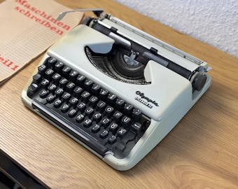 May include: A vintage Olympia Splendid 33 typewriter in a light green color, with black keys and a silver carriage return lever. A book with German text is visible next to the typewriter.