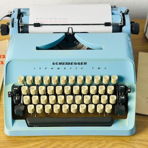 May include: A light blue typewriter with a black keyboard and the text "SCHEIDEGGER TVPOMATIC TMG" on the front. The typewriter has a white paper roll and a black carriage return lever.