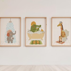 May include: Three framed art prints featuring cartoon animals in a bathroom setting. An elephant under a shower, a crocodile in a bathtub, and a giraffe on a toilet. Each print has a light wood frame and a neutral background.