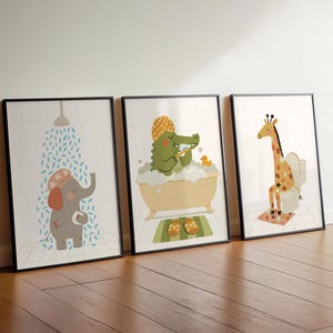 May include: Three framed art prints featuring cartoon animals in a bathroom setting. An elephant takes a shower, a crocodile bathes, and a giraffe sits on a toilet. The prints have black frames and are set against a white wall and wooden floor.