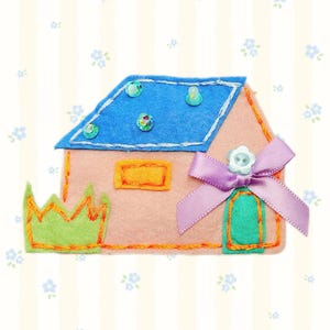Könnte beinhalten: Ein handgefertigtes Filzhaus mit blauem Dach, verziert mit Perlen. Das rosa Haus hat ein orangefarbenes Fenster, eine grüne Tür und ein lila Band mit einem Knopf. Links ist eine grüne Krone.