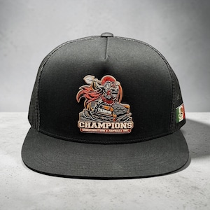 May include: Black trucker hat with a flat brim and mesh back. The front features a colorful patch with a gladiator graphic and the word "CHAMPIONS". A small Mexican flag patch is on the side.