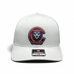 Peut inclure: Une casquette trucker blanche avec un logo brodé central représentant une tête d'ours bleu et rouge à l'intérieur d'un grand C, avec le texte CUBS en dessous. Un autocollant noir sur la visière affiche le texte RICHARDSON 112 ORIGINAL ADJUSTABLE.