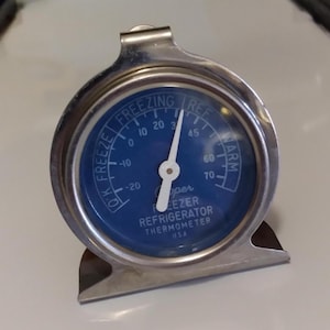 May include: A Cooper freezer refrigerator thermometer with a blue face and a silver metal casing. The thermometer has a needle that indicates the temperature, with markings for freezing and warm temperatures. The text "FREEZING" and "REF" are visible.