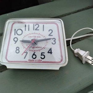 May include: A vintage white electric alarm clock with a red and white face. The clock face has black numbers and hands, with the words "LIGHTED DIAL" and "Ingraham" printed on it. The clock is marked "Made in USA".