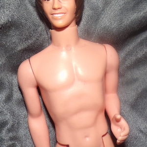 May include: A vintage doll with a tan complexion, brown hair, and a smiling expression. The doll is nude, with articulated joints and a defined physique. The doll's hair is styled in a classic, layered cut.