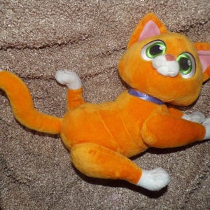 May include: Orange plush cat toy with white paws and tail tip. The cat has large green eyes, pink inner ears, and a purple collar. The toy is lying down on a textured surface.