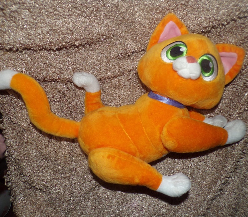 May include: Orange plush cat toy with white paws and tail tip. The cat has large green eyes, pink inner ears, and a purple collar. The toy is lying down on a textured surface.