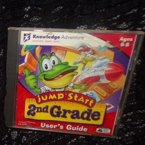 May include: Jump Start 2nd Grade CD-ROM in a clear plastic case. The cover features a cartoon frog, a rocket ship, and the text "Jump Start 2nd Grade" in yellow and red. The case is labeled "User's Guide" and "Ages 6-8".