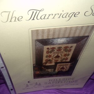 May include: A cream-colored paper with the title "The Marriage Sampler" printed in black script. The paper features a framed cross-stitch sampler with the text "Our happiness is in the day that we were blessed by the Lord." The sampler is surrounded by a brown frame and a striped background. The text "Hillside Samplings" is printed at the bottom of the paper.